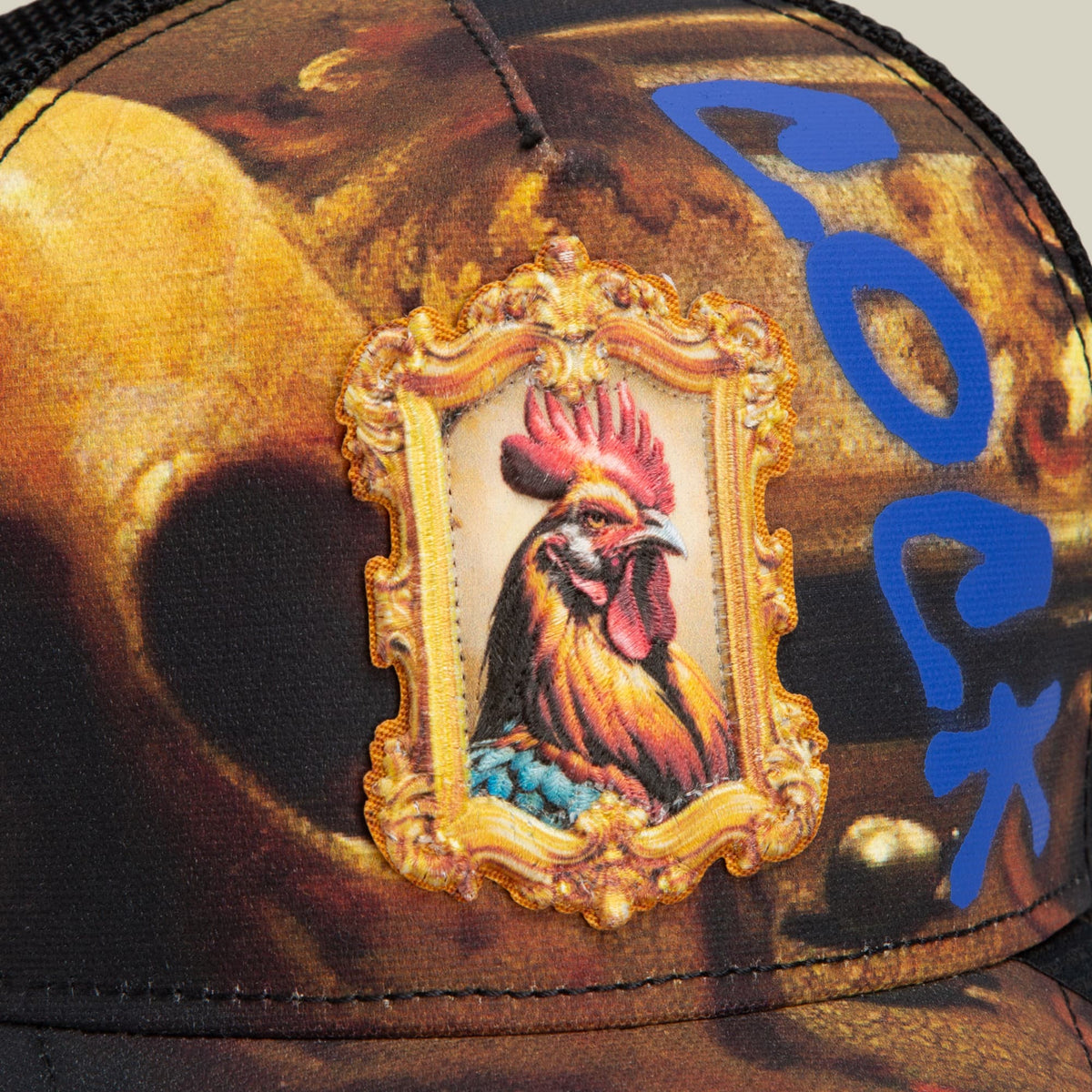 The Penitent Magdalene by Goorin Bros is a brown trucker hat featuring ornate gold framing with rooster embroidery in red and gold tones, accented with blue decorative elements on the side.