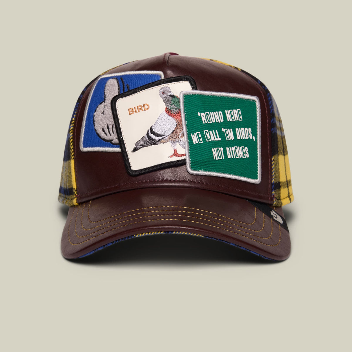 The Bird! by Goorin Bros. is a brown trucker hat featuring embroidered bird and text patches on the front, with yellow and navy plaid fabric on the mesh panels, displayed against a neutral background.