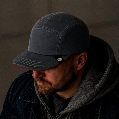 A man wearing Goorin Bros. North Boulder, a hand cut and sewn 5-panel camp cap, along with a hooded sweatshirt and denim jacket, looks down in a dimly lit setting.