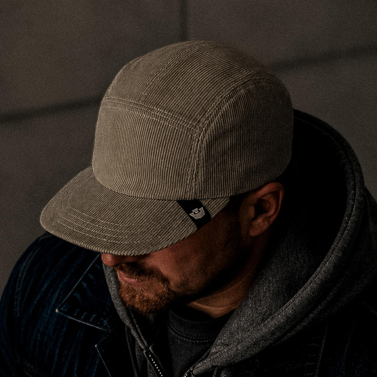 A man in low lighting wears the Goorin Bros. Meadow Creek, a handmade in USA beige corduroy cap, along with a gray hoodie and dark jacket, facing downward.