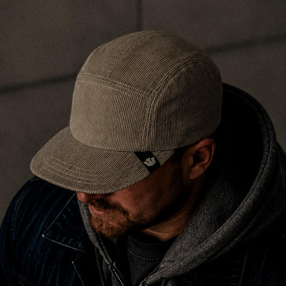 A man in low lighting wears the Goorin Bros. Meadow Creek, a handmade in USA beige corduroy cap, along with a gray hoodie and dark jacket, facing downward.