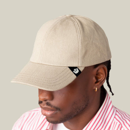 A person exudes subtle confidence wearing the Goorin Bros. Noisy Tailored Baseball cap in beige, featuring a small black logo tag, paired with a pink-and-white striped shirt against a plain light background.