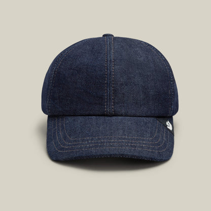 The Noisy Tailored Baseball cap by Goorin Bros. is featured in indigo denim with visible stitching, set against a plain beige background to emphasize its color-driven style and understated confidence.