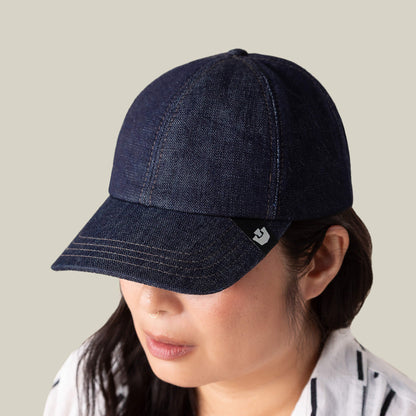 Wearing the "Noisy Tailored Baseball" cap by Goorin Bros.—a dark denim hat featuring a subtle black and white logo tag—against a beige backdrop, this look exudes understated confidence and color-driven style.
