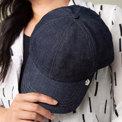 A person holds the Noisy Tailored Baseball cap by Goorin Bros. in dark blue denim, paired with a white shirt featuring black stripes, embodying subtle confidence and color-driven style.