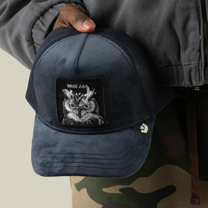 A person in a gray jacket and camo pants holds the Goorin Bros. Velour Wise Ass trucker hat, a navy blue mesh-back cap featuring an owl patch and the text WISE ASS on the front.