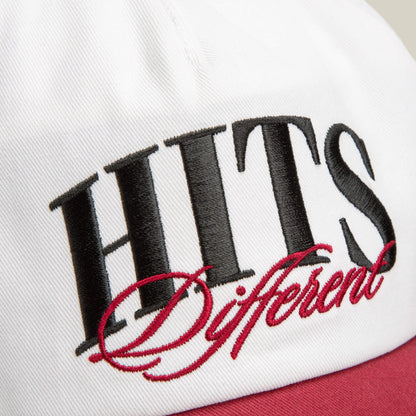 Close-up of the Goorin Bros. Hits Different cap in white and maroon, featuring oversized "HITS Different" embroidery in black and red on the front and a curved brim.