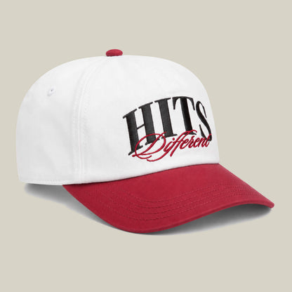 The Goorin Bros. Hits Different cap is white with a red curved brim and button, featuring "HITS Different" embroidered in black and red script on the front.
