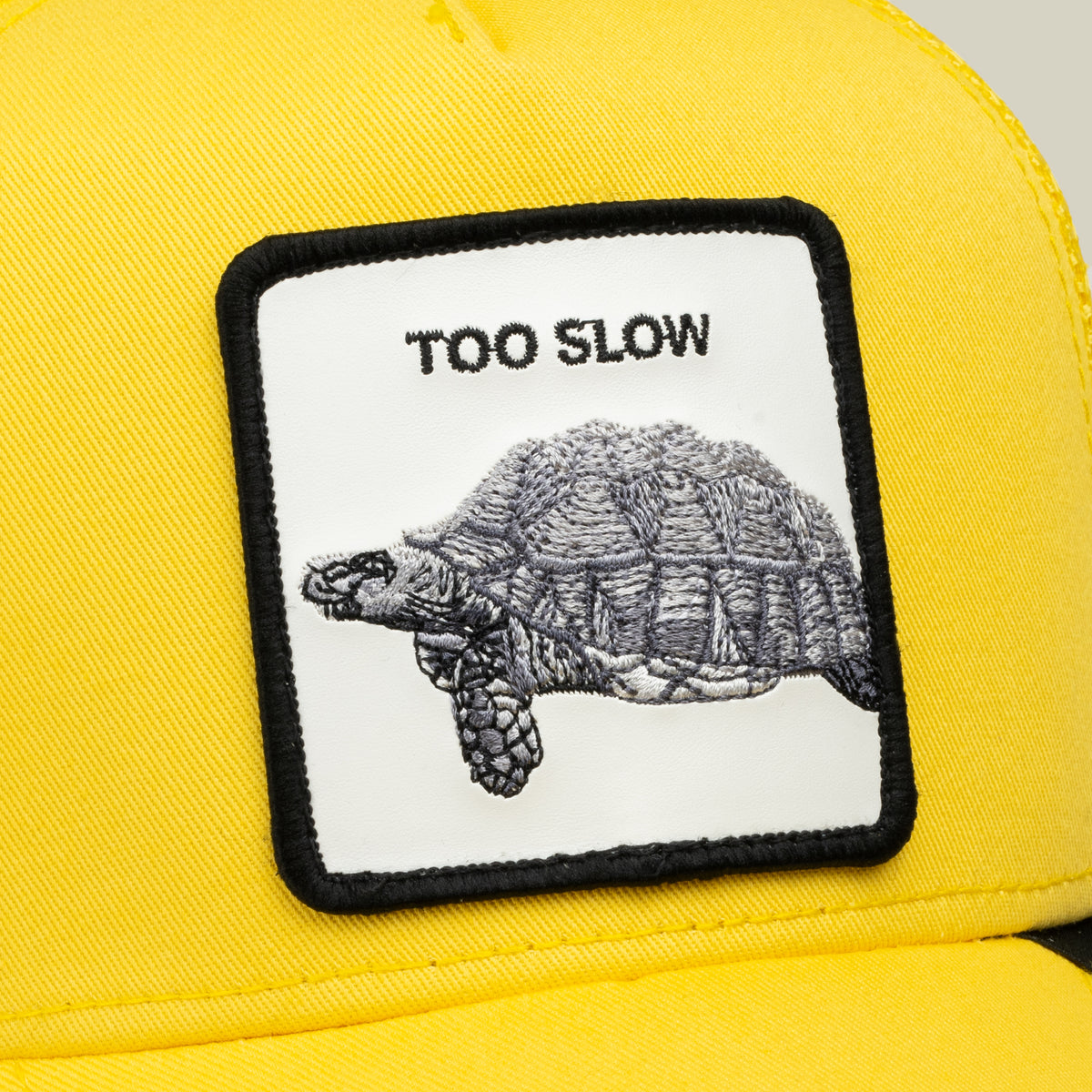 Close-up of the Goorin Bros. “Down Low...” yellow cap, featuring a rectangular patch with an embroidered tortoise and the words TOO SLOW above it—a true badge of honor for those lucky enough to snag this elusive drop.