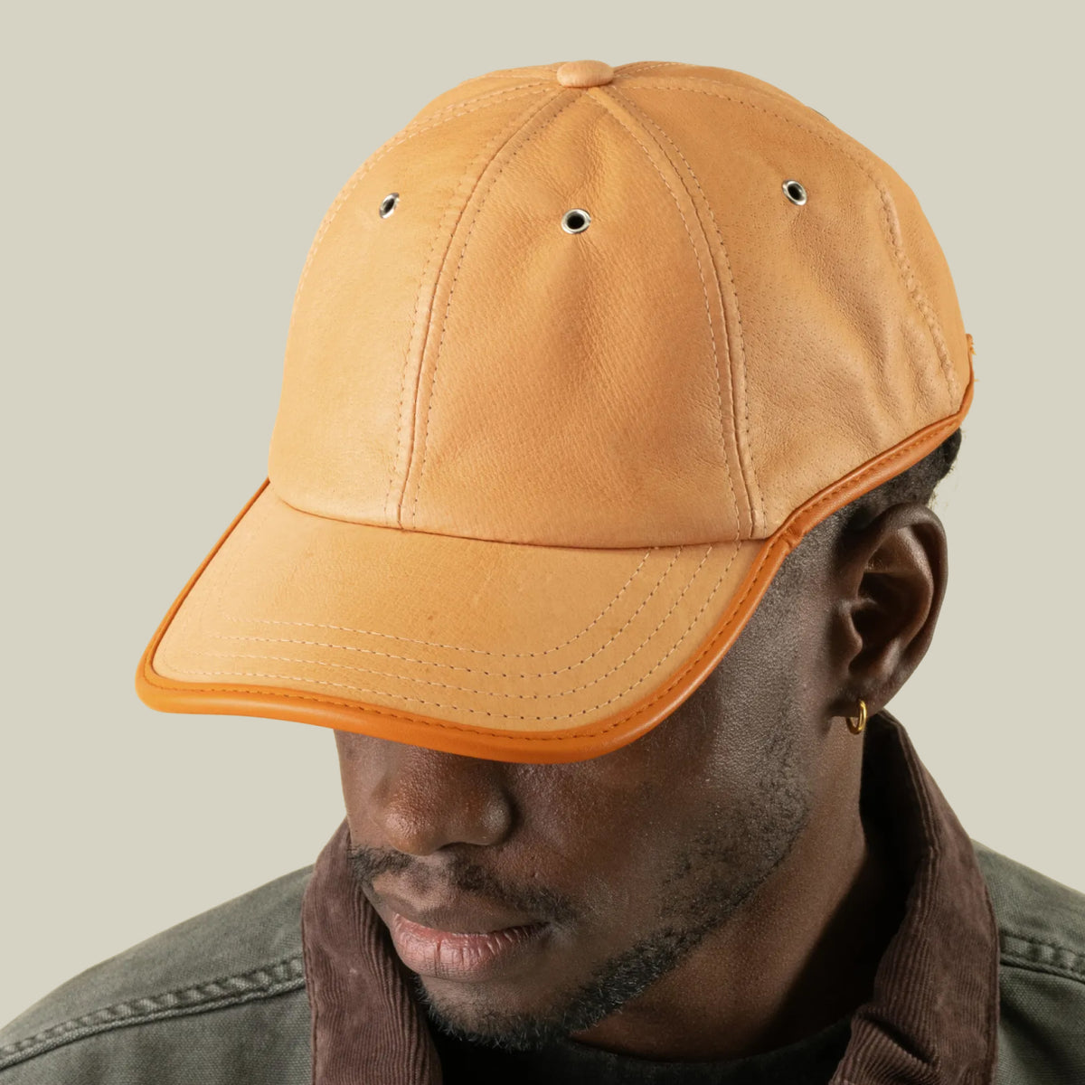 A person wearing the Goorin Bros. Steiner tan suede papa cap with orange trim and an adjustable plaid strap, looking down against a plain light beige background.