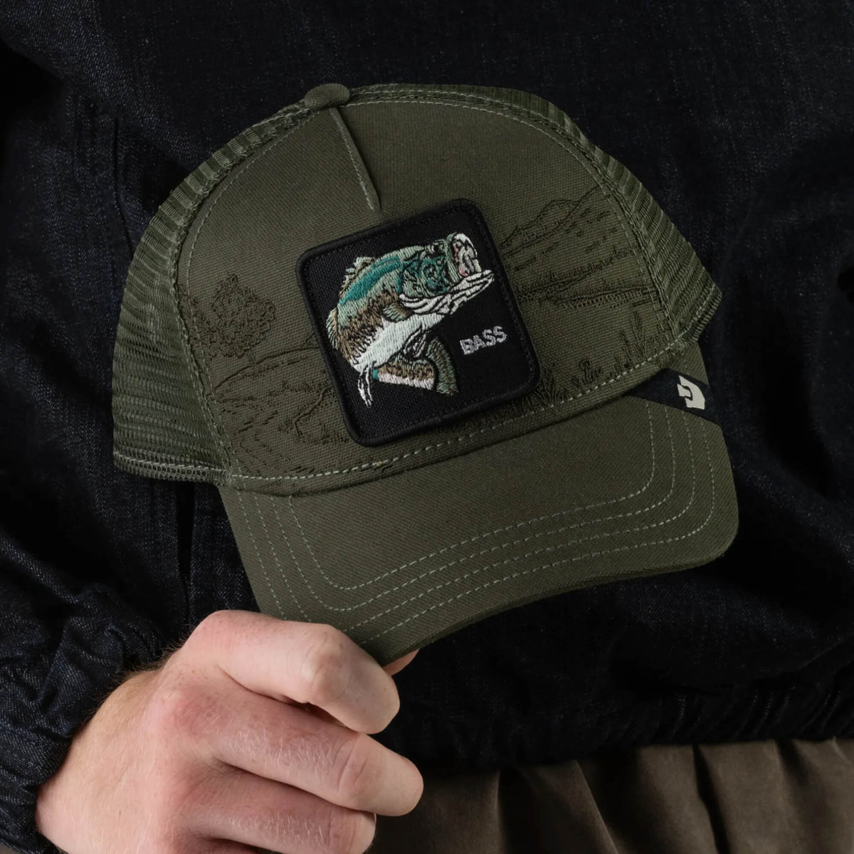 A person holds the Badlands Bass trucker hat by Goorin Bros., featuring an olive green design with a patch of a jumping bass fish and “BASS” embroidered below. This breathable mesh hat is perfect for fishing enthusiasts.