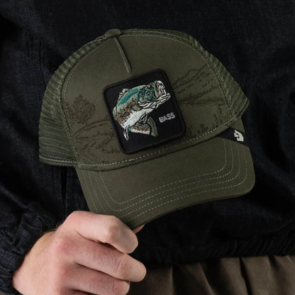 A person holds the Badlands Bass trucker hat by Goorin Bros., featuring an olive green design with a patch of a jumping bass fish and “BASS” embroidered below. This breathable mesh hat is perfect for fishing enthusiasts.