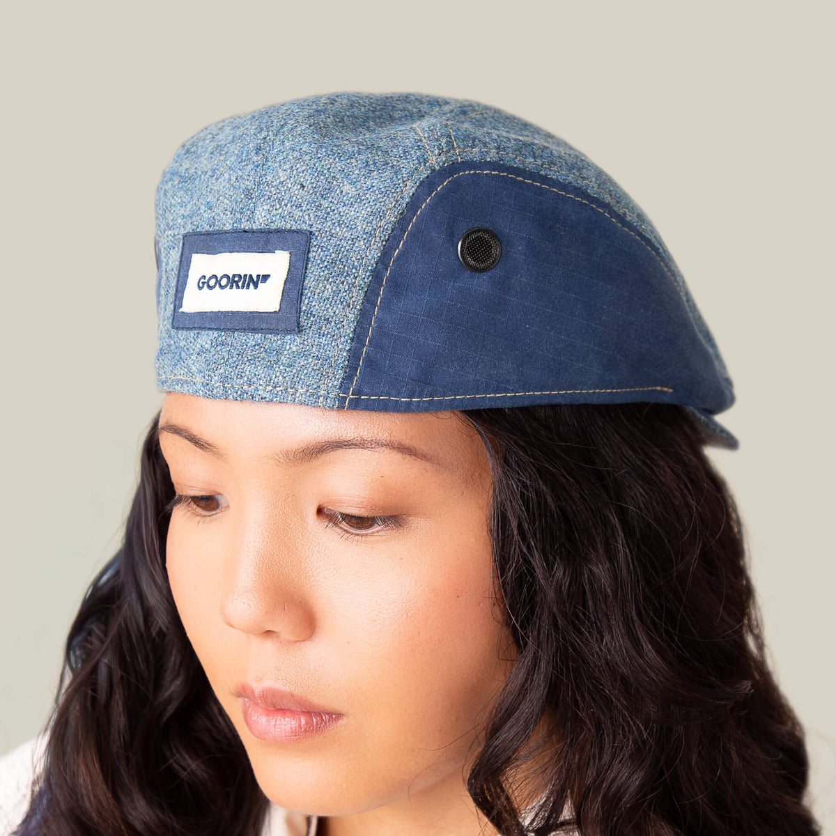 Someone wearing the Full Wattage blue tweed flatcap from Goorin Bros., featuring a label on the front, looks downward against a plain background.