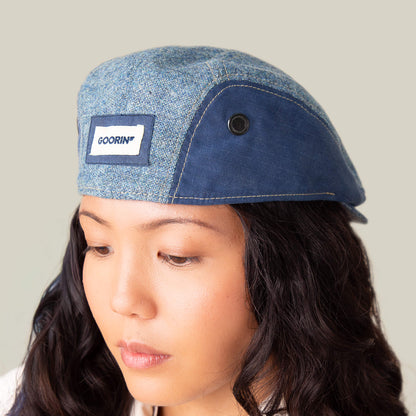 Someone wearing the Full Wattage blue tweed flatcap from Goorin Bros., featuring a label on the front, looks downward against a plain background.