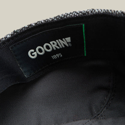 Close-up of the inside label on a grey tweed cap, showing the brand "Goorin Bros." and the product name "Gibbins" on a black fabric background—a classic piece for men's fashion.