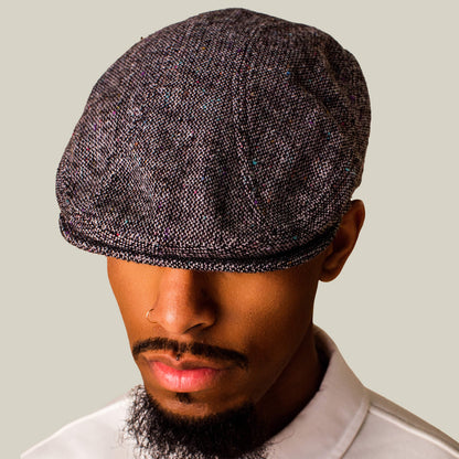 A man wearing the Gibbins textured grey tweed cap by Goorin Bros., a classic white collared shirt, and a nose ring stands against a plain background, his eyes partially hidden by the flatcap—a stylish touch to men's fashion.