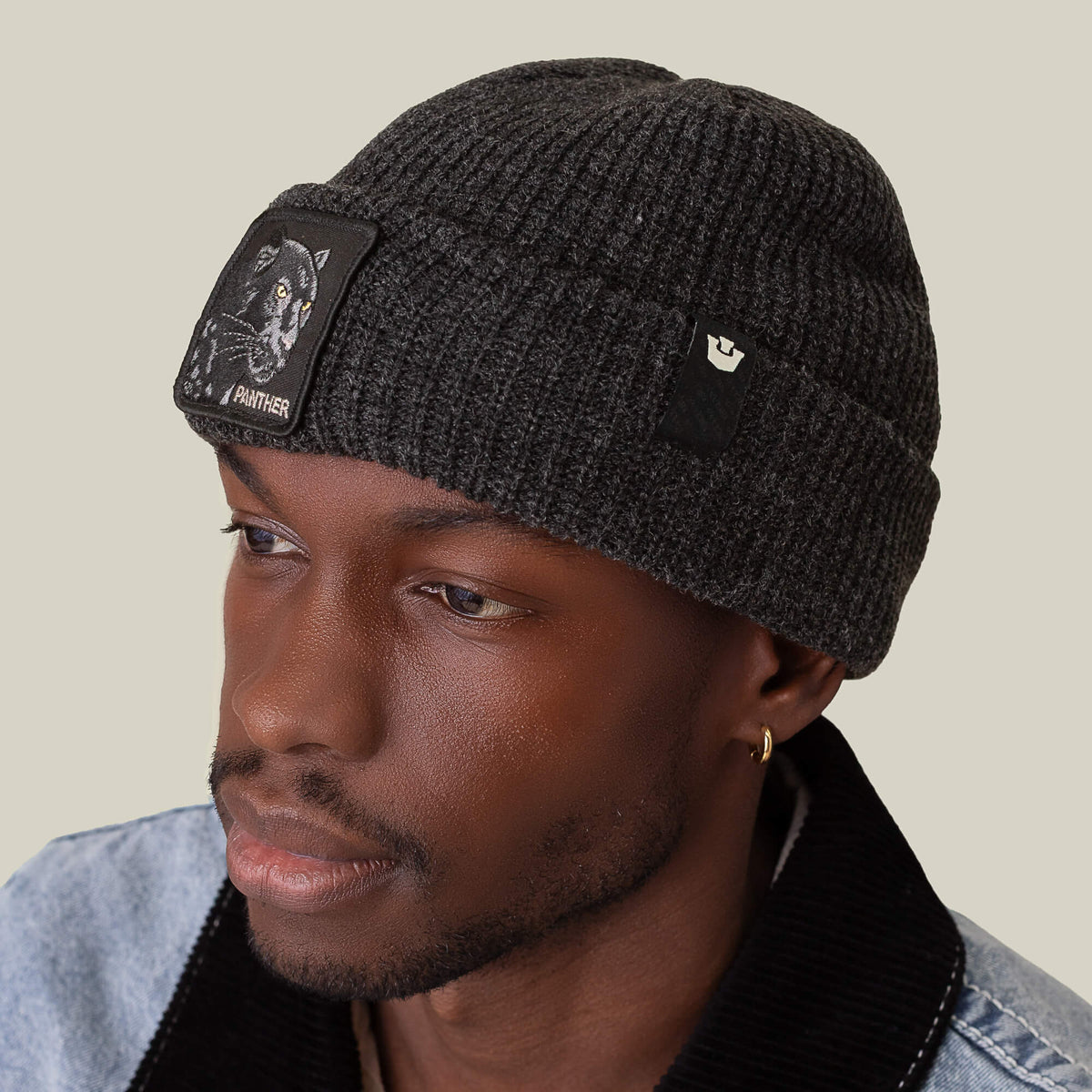 A man in Goorin Bros.'s The Panther Beanie, featuring two patches and a dark gray design, pairs it with a light denim jacket as he looks to the side against a plain background.