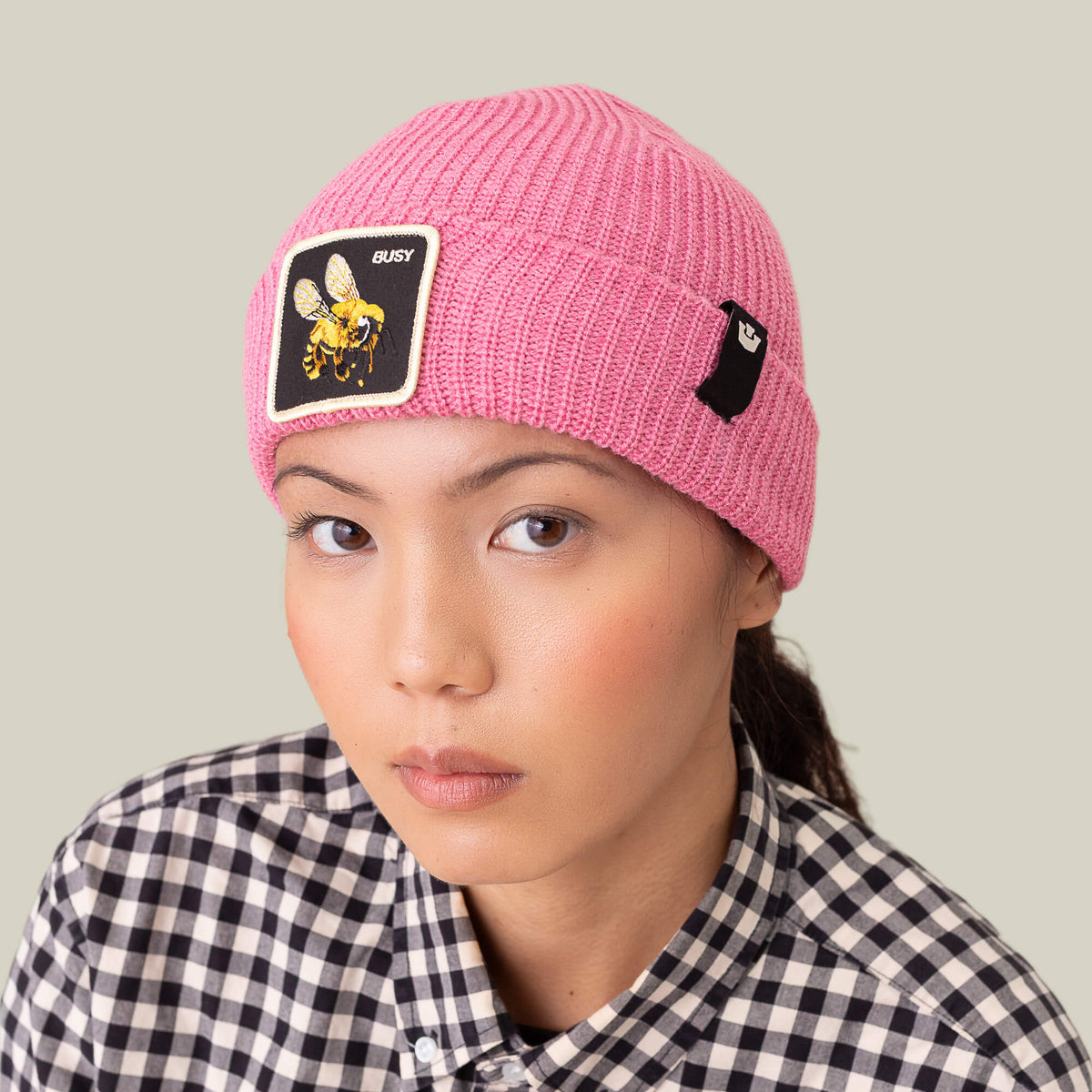 A person models The Busy Beanie by Goorin Bros.—a vibrant pop-pink knit hat with a buzzing bee patch—paired with a black-and-white checkered shirt, gazing at the camera against a plain backdrop.