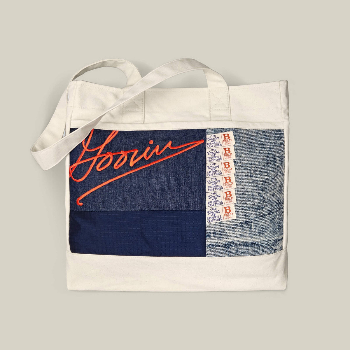 The Goorin Bros. Goorin Reworked Tote in EDGE features a white base with blue patchwork, red cursive Goorin text, and a vertical row of small red and white B tags on the front.
