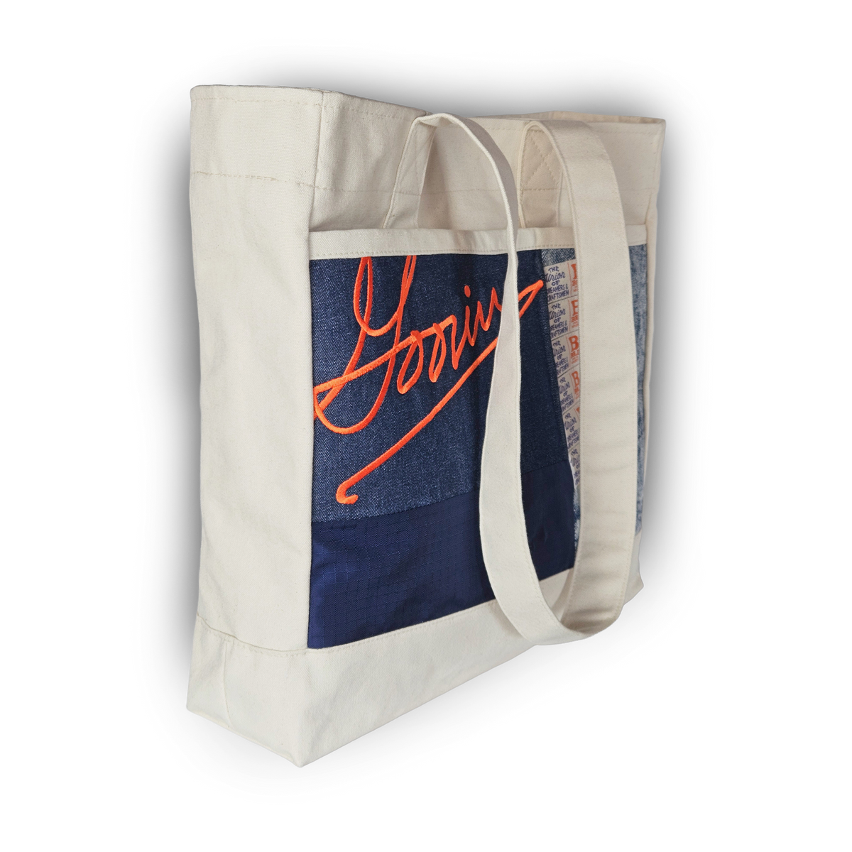 The Goorin Bros. Goorin Reworked Tote is a white canvas bag with long handles, a blue patch, and orange “Goorin” script. Made in the USA from upcycled fabric for an eco-friendly, stylish look.
