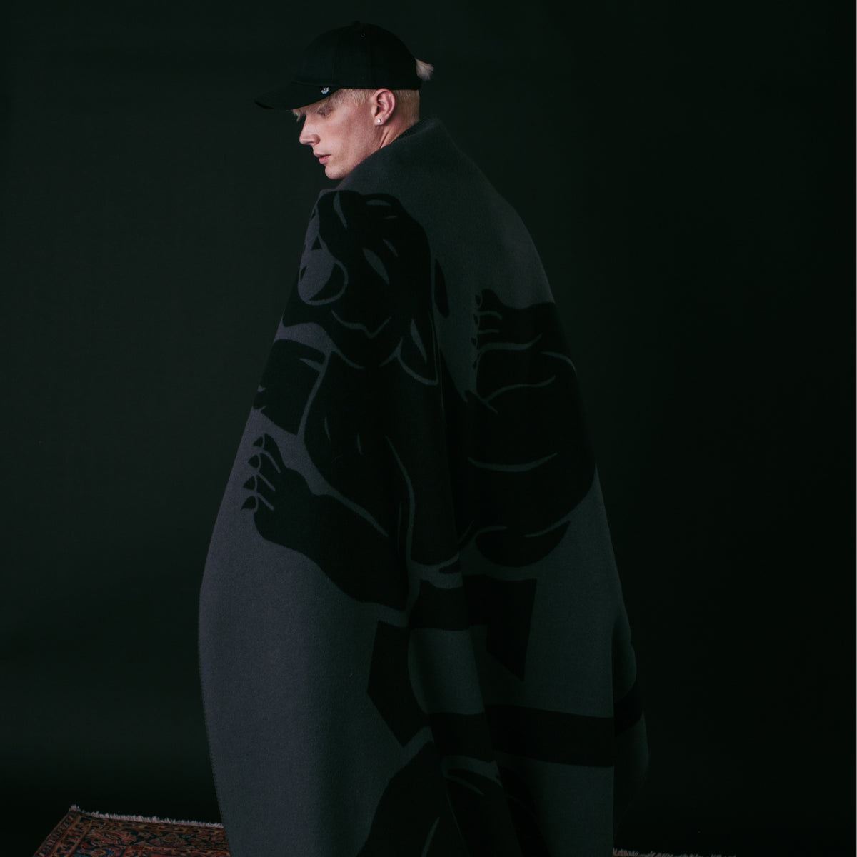 A person stands against a dark background, draped in the Goorin Bros. Panther Crest Blanket featuring a bold graphic design, and wearing a black cap.