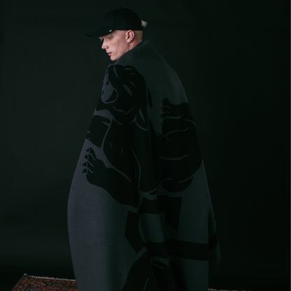 A person stands against a dark background, draped in the Goorin Bros. Panther Crest Blanket featuring a bold graphic design, and wearing a black cap.
