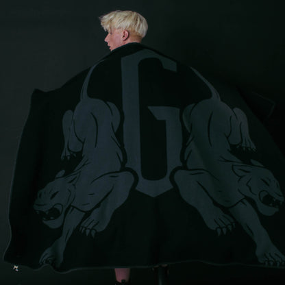 A person with short blond hair is wrapped in a Goorin Bros. Panther Crest Blanket, which features a large “G” and two stylized panthers on a solid black background.