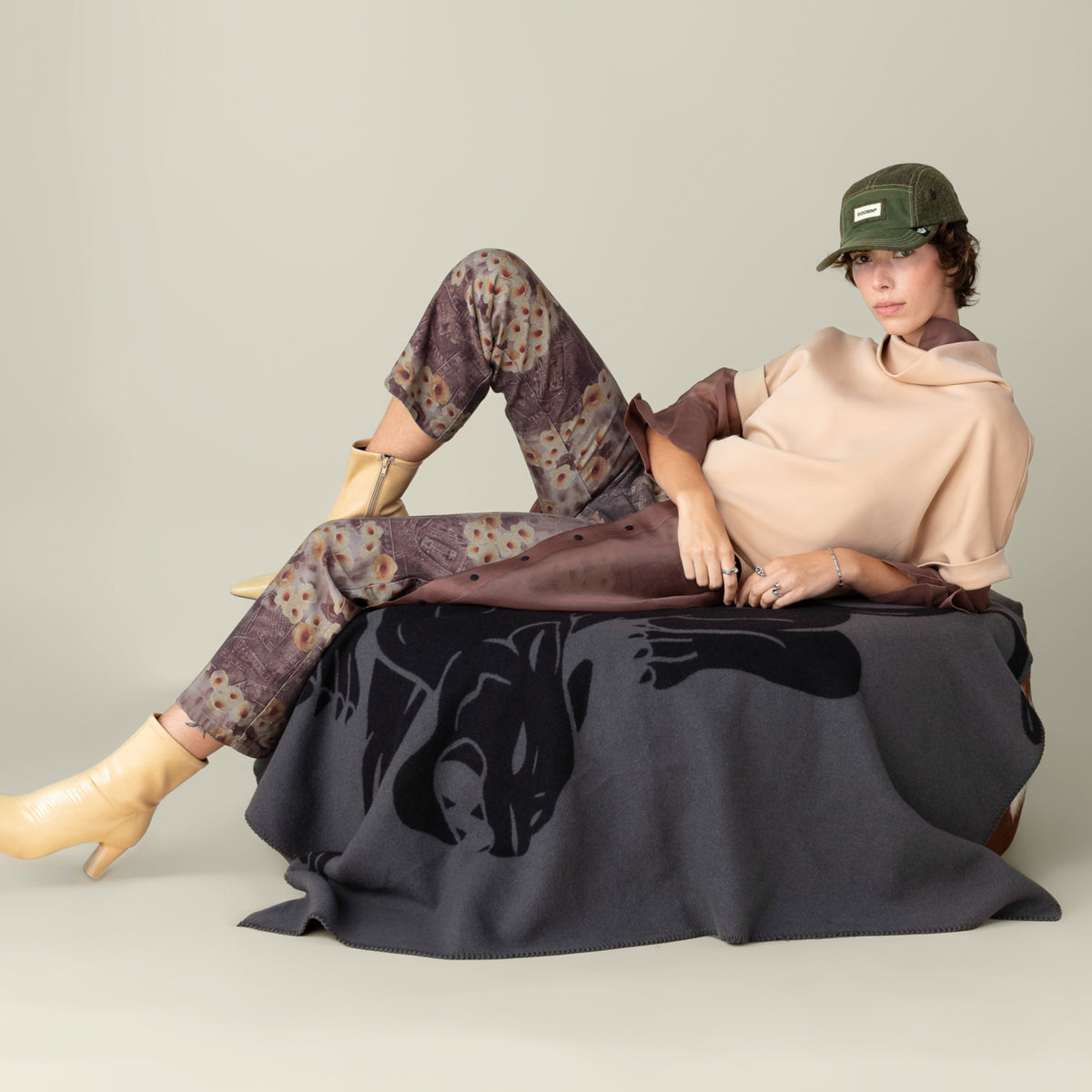 A person in a beige top, patterned pants, tan boots, and a green cap sits on the Goorin Bros. Panther Crest Blanket featuring a bold panther design, set against a plain beige background.