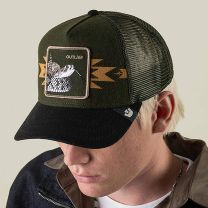 A person wearing the Goorin Bros. Canyon Outlaw hat—a green and black wool blend trucker featuring a rattlesnake patch labeled OUTLAW and geometric side designs—channels pure outlaw energy.