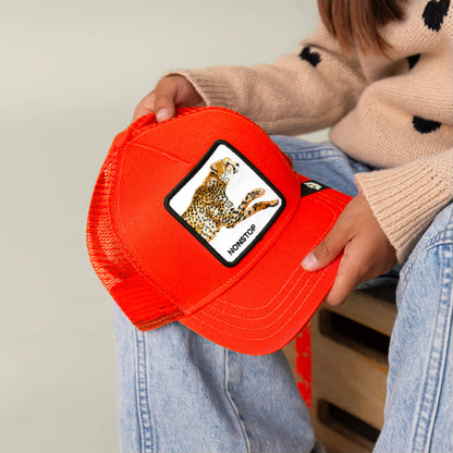 A child holds The Nonstop Cheetah Mini Noir cap by Goorin Bros., featuring an orange mesh design and a cheetah patch with the word NONSTOP. Paired with a beige sweater adorned with black spots and blue jeans, the vibrant orange crown of the cap adds a playful accent to their look.