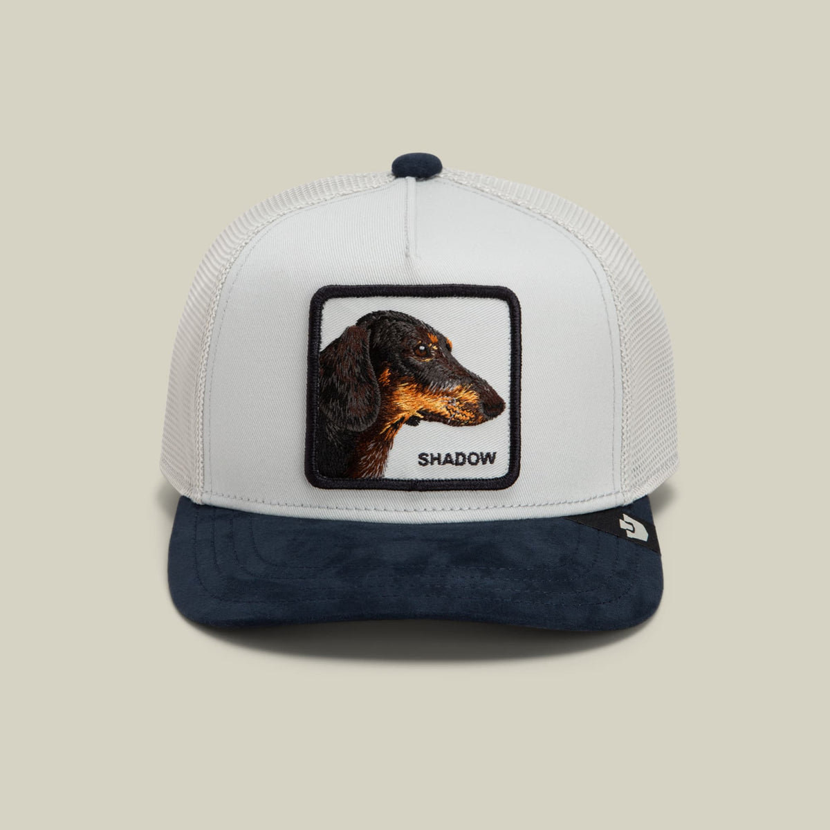 The Goorin Bros. Suede Shadow Mini is a white and navy trucker hat featuring a mesh back and embroidered dachshund patch with text reading "Shadow" on the front.