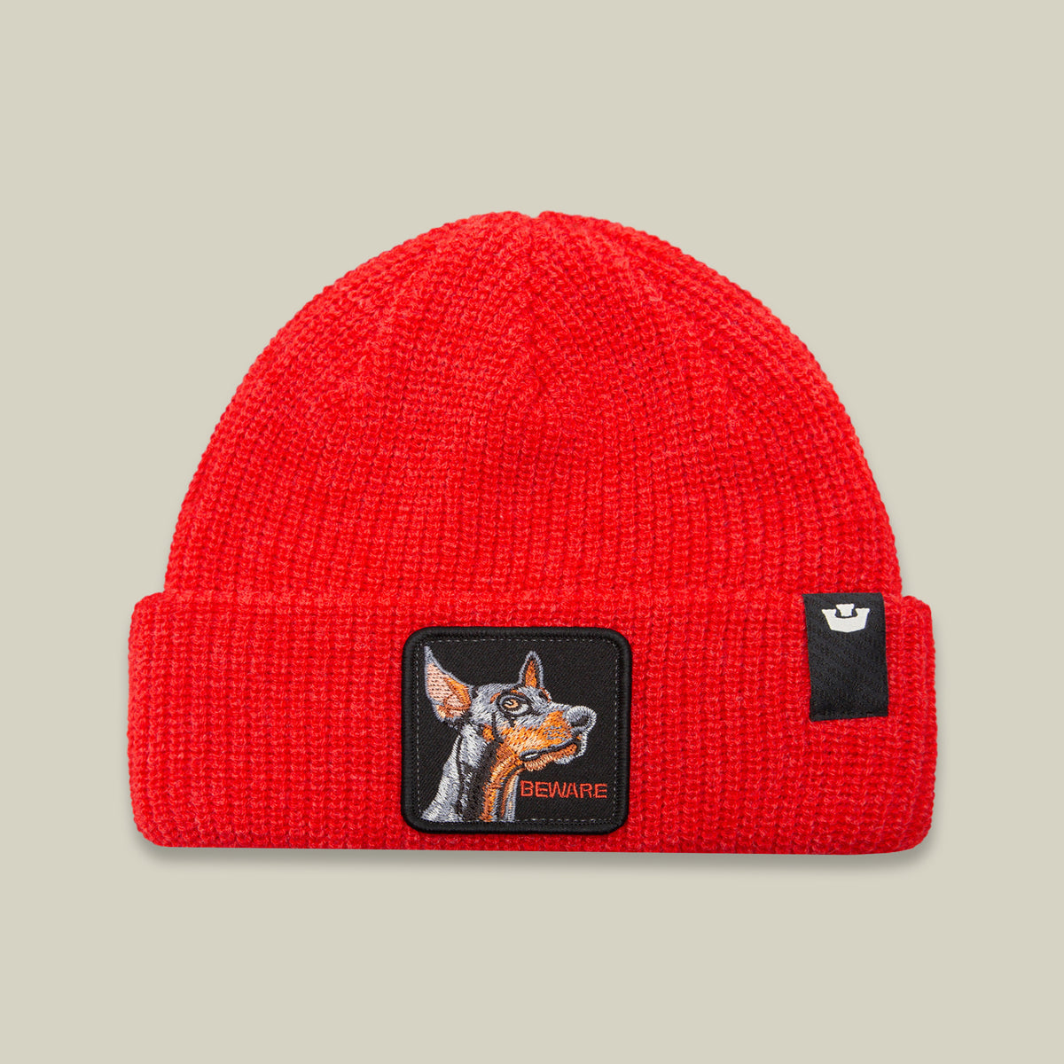 The Beware Dog Beanie by Goorin Bros. is a red heavyweight knit hat featuring a dog patch and the word “BEWARE” embroidered on the front, displayed on a beige background. This bold beanie brings warmth and attitude to your winter wardrobe.