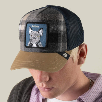 A man wearing the Brit Frenchie cap by Goorin Bros.—a stylish plaid adjustable hat with an embroidered mesh back, brown brim, and a patch featuring a dog illustration and the word “Dodgy” above it—perfect for any casual outing.