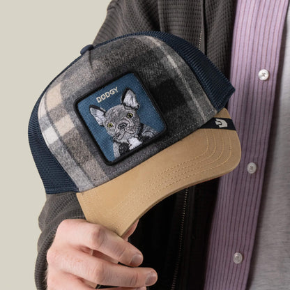 A person holds the Brit Frenchie cap by Goorin Bros., a plaid adjustable hat featuring a patch with an embroidered French Bulldog and the word “DOGGY,” making this playful and stylish accessory perfect for any dog lover.