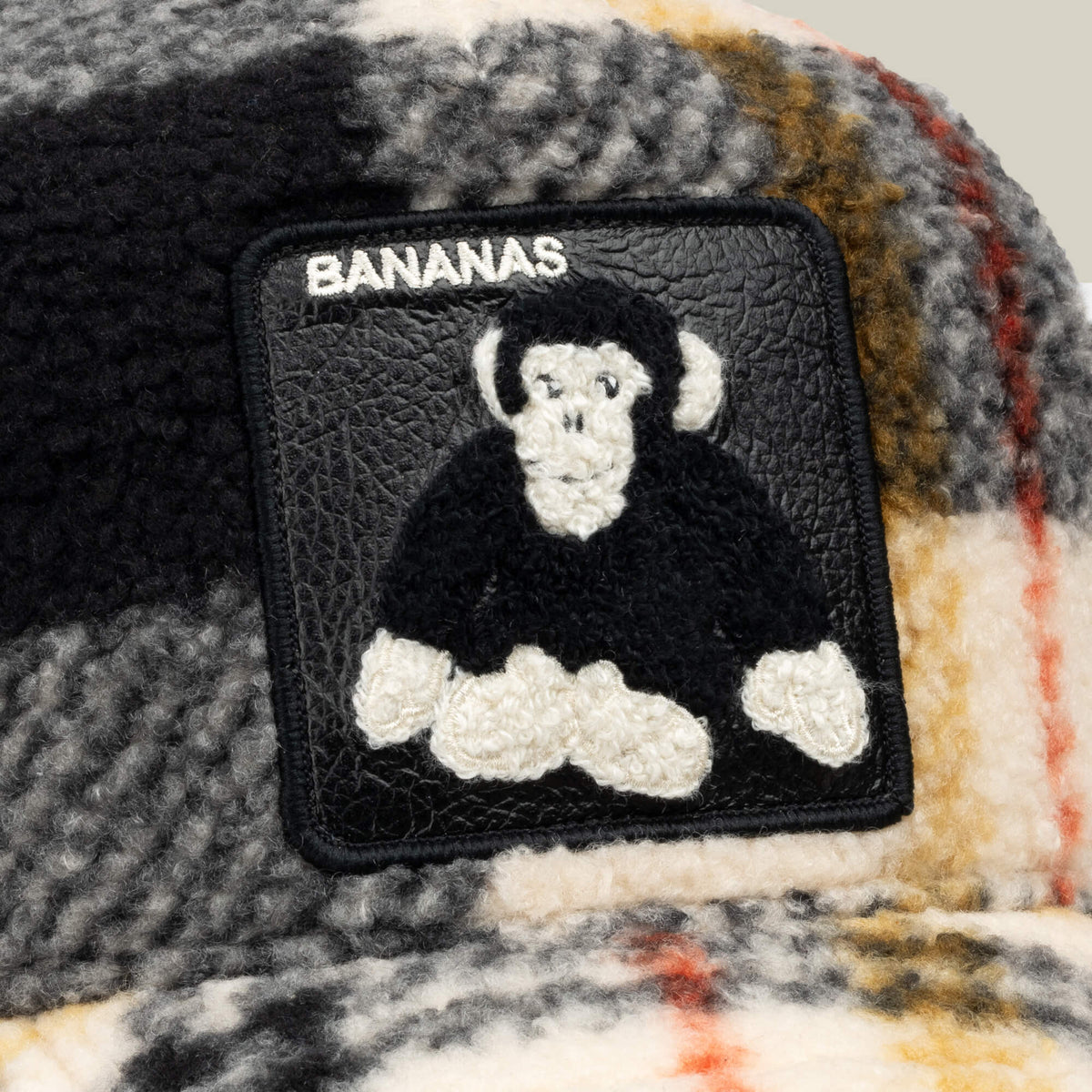 A playful patch featuring a cartoonish black and white monkey and the word “BANANAS” is sewn onto a plaid fuzzy fabric in black, white, yellow, and orange tones—making the Goorin Bros. Double Stuffed - Bananas hat a fun way to add quirky charm to your favorite fleece trucker or adjustable snapback.