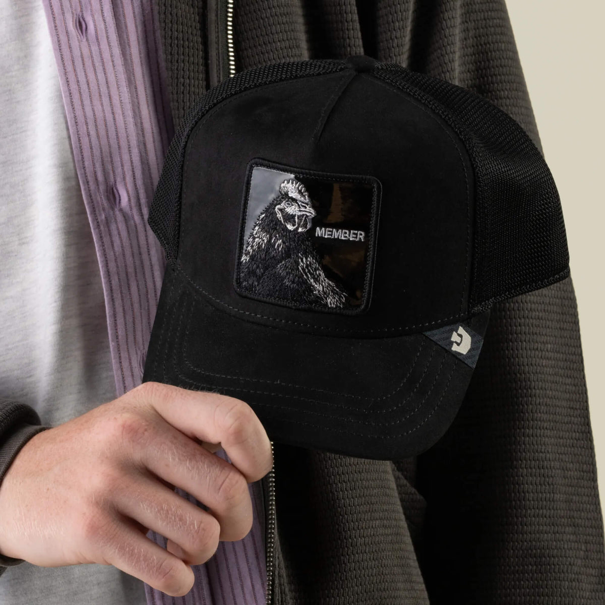 A person holds a jet-black polyester mesh baseball cap, the Black Out Rooster by Goorin Bros., featuring a MEMBER patch with a gorilla illustration on the front.
