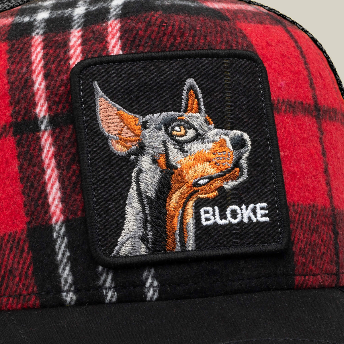 The Goorin Bros. Brit Bloke is a red-and-black plaid trucker hat featuring a black embroidered patch with an image of a Doberman and the word “BLOKE” in white text.