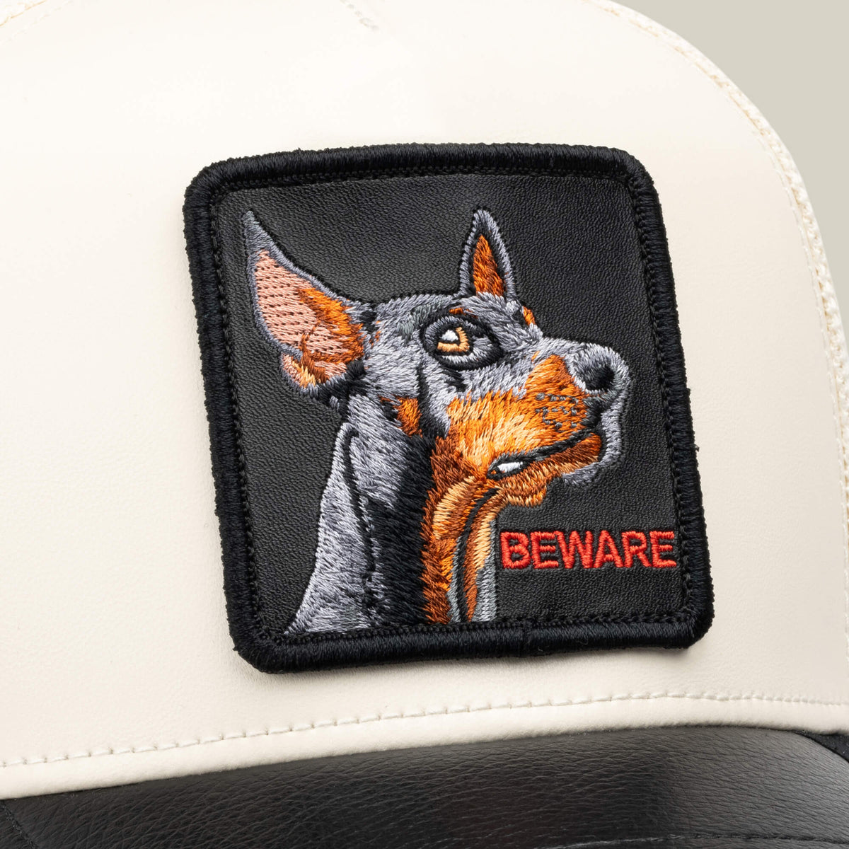 Close-up of the Goorin Bros. “Faux Leather Doberman” collectible hat, featuring a sewn patch with an embroidered dog and the word BEWARE in red letters on a white and black design.