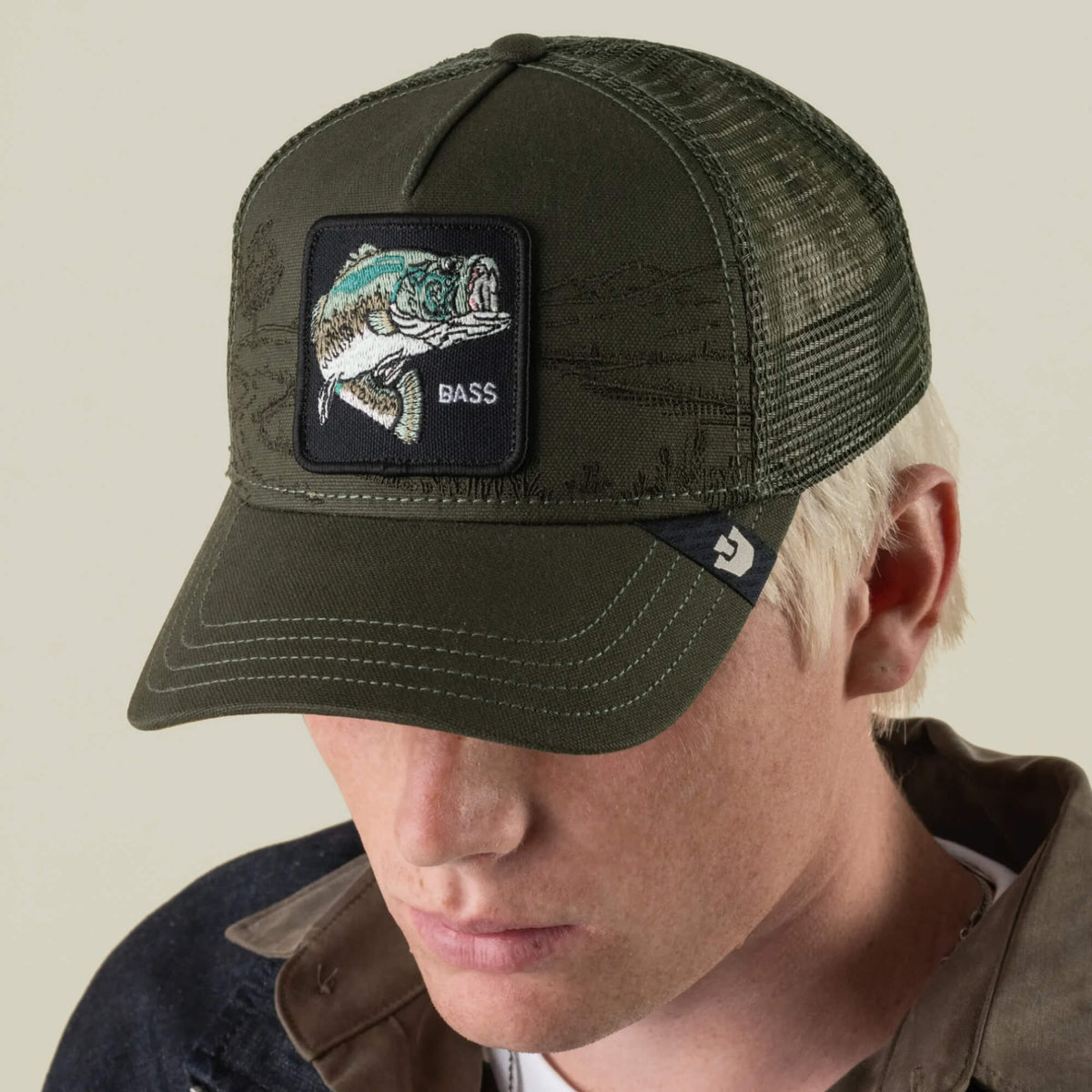 A person is wearing the Goorin Bros. Badlands Bass trucker hat, an olive green cap featuring a patch with an embroidered fish and the word “BASS.” With its breathable mesh design, this hat is perfect for outdoor adventures, as the wearer looks downward.
