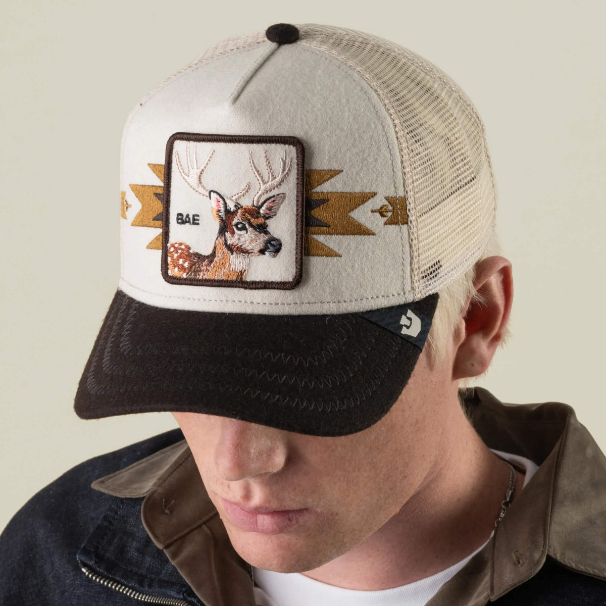 A person wearing the Goorin Bros. Canyon Bae cap, a tan and black mesh back hat with a patch featuring a deer and the word “BAE” embroidered on the front.