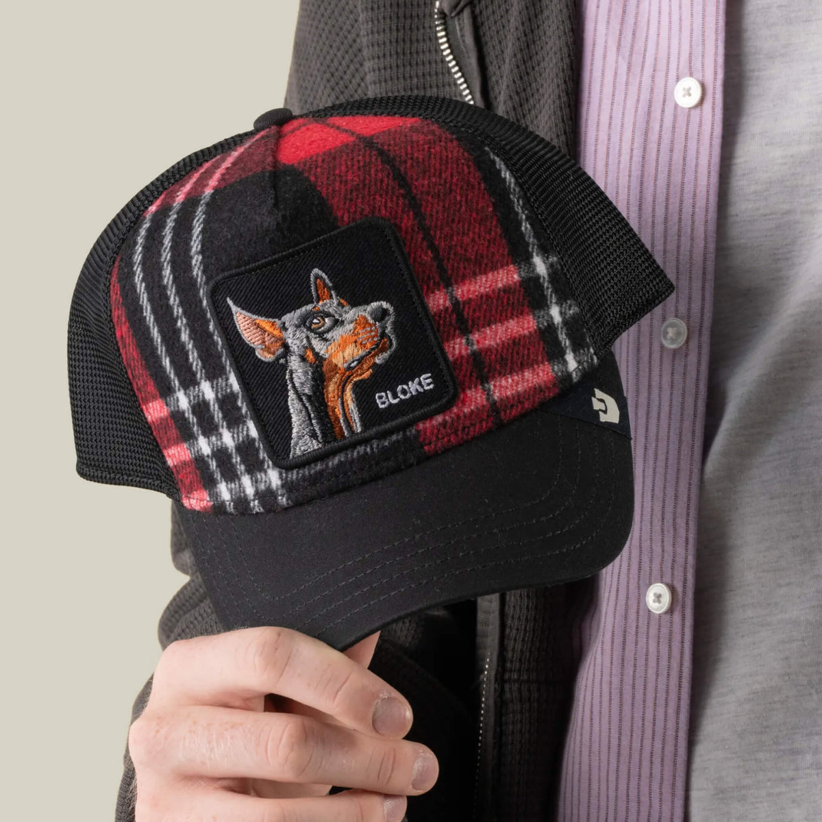 A person holding the “Brit Bloke” trucker hat by Goorin Bros., featuring a red-and-black plaid design, mesh back, and an embroidered BLOKE patch with a dog’s head.