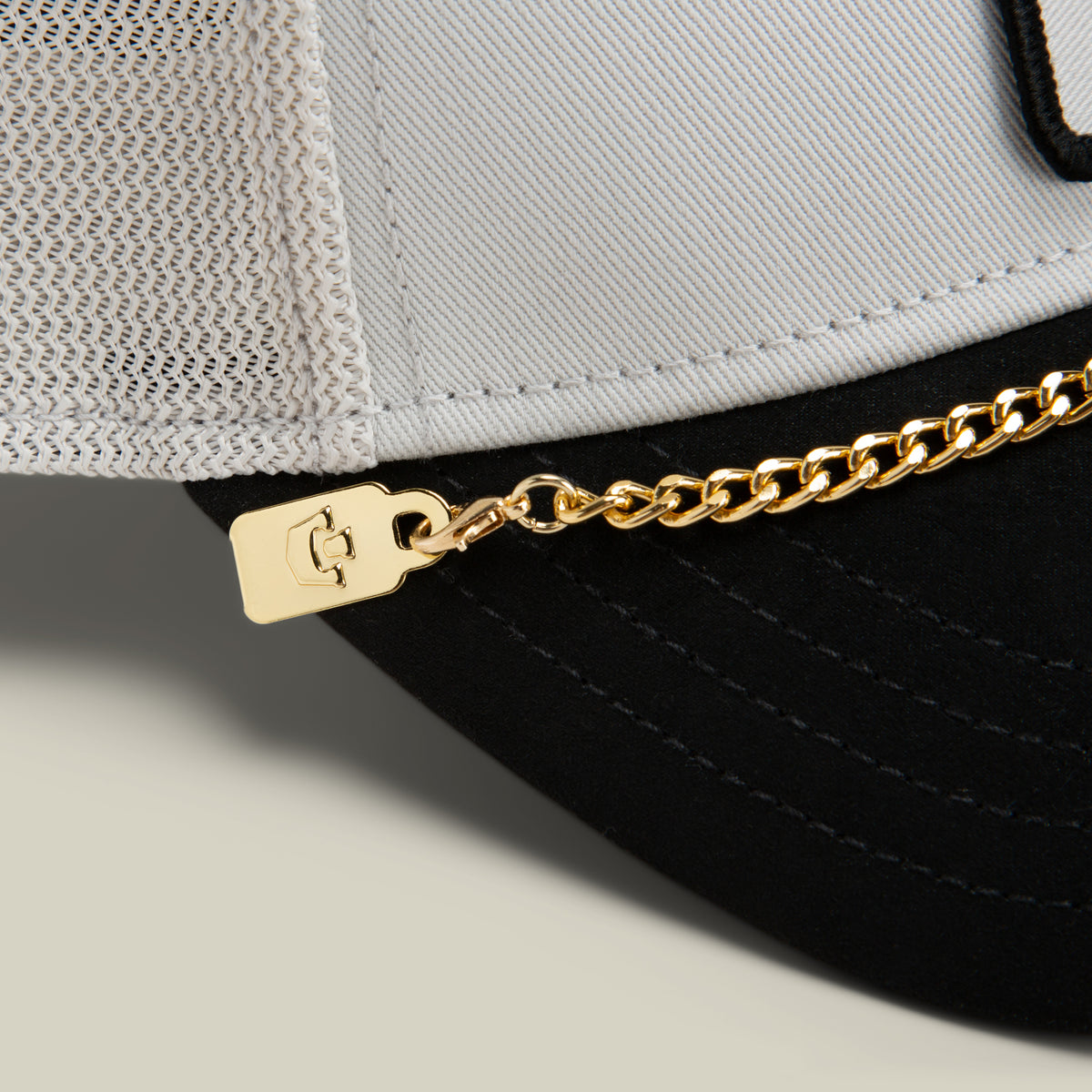 Close-up of a two-tone cap featuring the Goorin Bros. Mini CapLoc Chain—a striking piece of hat jewelry attached with a small metal tag—accented by a mesh back and black brim.
