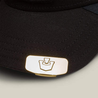Close-up of the Goorin Bros. Castle CapLoc— a black hat accented with a gold zinc alloy metal emblem showcasing a stamped geometric logo on the brim, designed as a stylish accessory.
