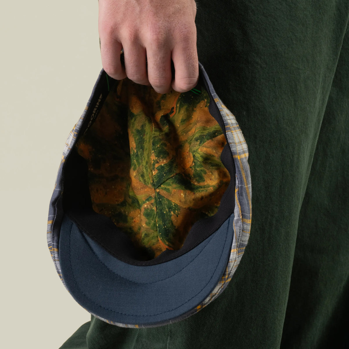 A person holding the Point Lobos 6 Panel Ivy cap by Goorin Bros. by its brim displays the inner lining, which features a green and orange leaf pattern, against a light background. Handmade in the USA from a breathable cotton blend, this blue plaid cap offers all-day comfort and style.