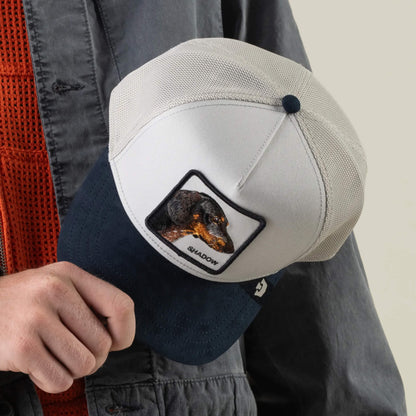 A person holds the Goorin Bros. Suede Shadow hat, a two-tone trucker cap featuring a rectangular patch with an embroidered dog and the word “SHADOW” beneath it, finished with a stylish suede brim.