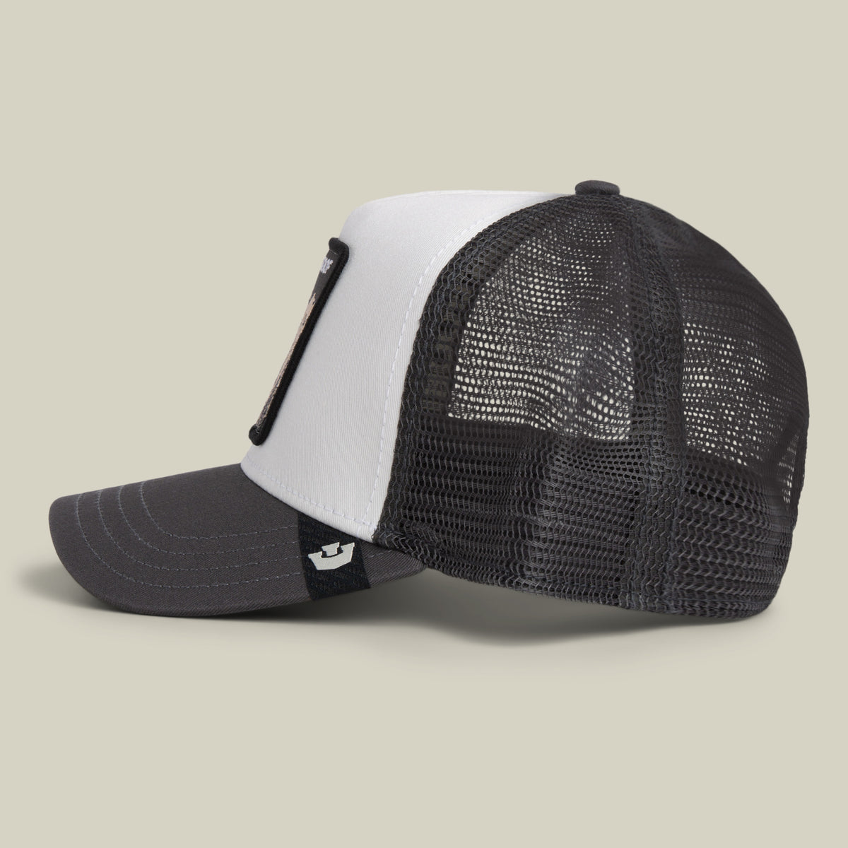 Side view of the Goorin Bros. Bulletproof Trucker hat, featuring a black and white mesh back design with a curved brim and a black embroidered patch on the front panel, displayed against a plain beige background.