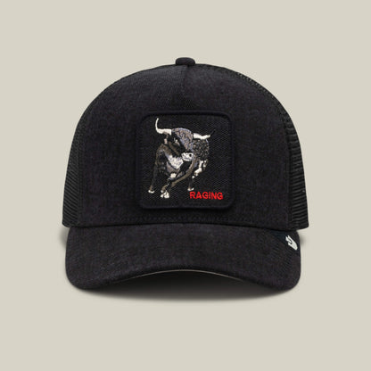 The Goorin Bros. Denim Raging is a black denim trucker hat featuring a front patch with an embroidered bull and the word “RAGING” in bold red text. This horned patch cap combines a classic style with breathable mesh trucker panels, all displayed against a simple beige background.