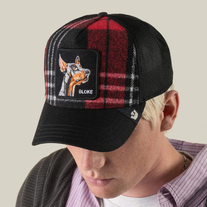 A man with light hair is looking downward while wearing the Goorin Bros. ‘Brit Bloke’ trucker hat, which features a red-and-black plaid pattern and an embroidered BLOKE patch with a cartoon Doberman.