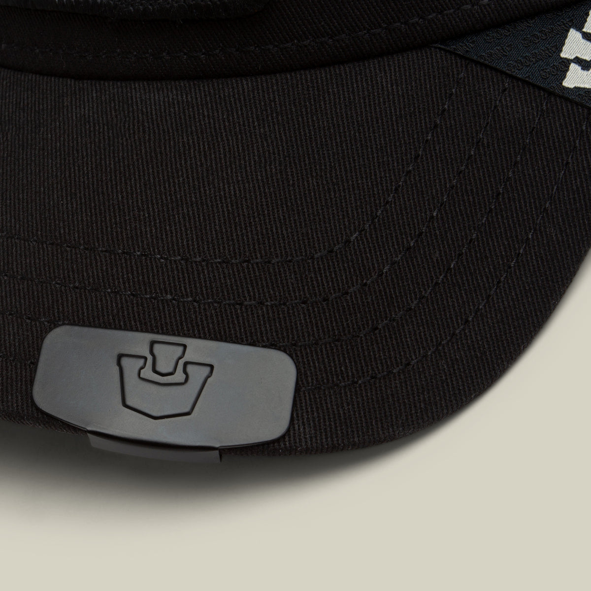 Close-up of a black cap by Goorin Bros., featuring the Castle CapLoc—a small, gray hat accessory made from durable zinc alloy—clipped to the brim, with a partial white logo visible on the side.