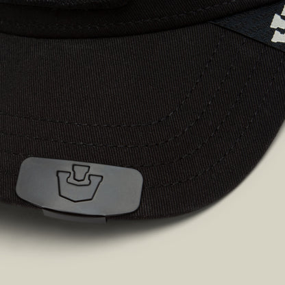 Close-up of a black cap by Goorin Bros., featuring the Castle CapLoc—a small, gray hat accessory made from durable zinc alloy—clipped to the brim, with a partial white logo visible on the side.